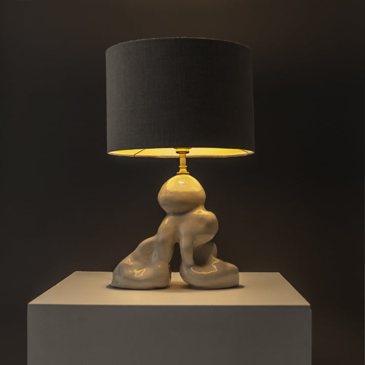 Designer stoneware lamp glowing warmly under a linen shade – ideal for cozy corners and evening ambience. / Dizainerio akmens masės šviestuvas su lininiu gaubtu skleidžia šiltą šviesą – jaukioms namų erdvėms.