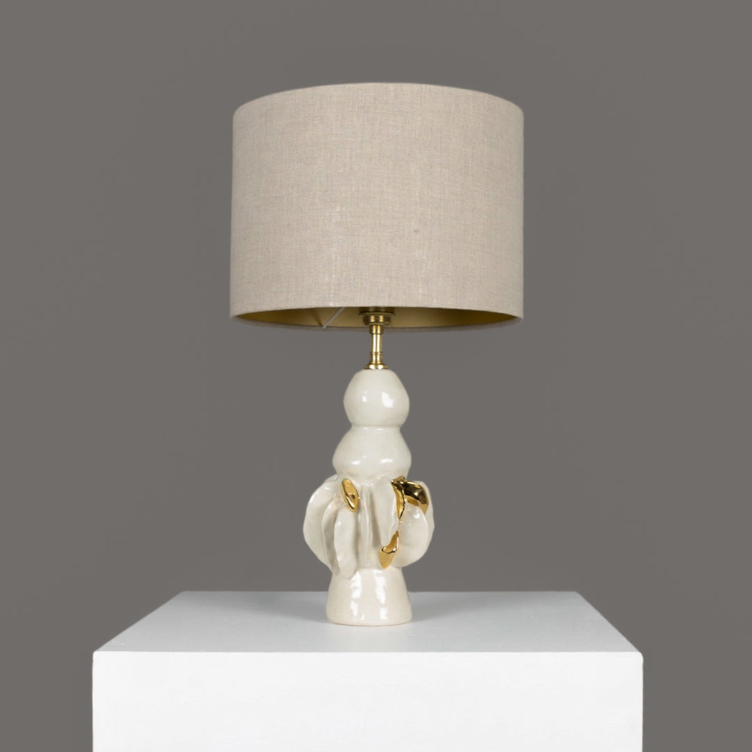 Sculptural ceramic table lamp with 100% linen beige lampshade and gold detail – elegant lighting for modern art-filled homes. / Skulptūrinis keraminis stalinis šviestuvas su 100 % lino smėlio spalvos gaubtu ir aukso detalėmis – moderniam, meniškam interjerui.