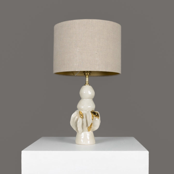 Sculptural ceramic table lamp with 100% linen beige lampshade and gold detail – elegant lighting for modern art-filled homes. / Skulptūrinis keraminis stalinis šviestuvas su 100 % lino smėlio spalvos gaubtu ir aukso detalėmis – moderniam, meniškam interjerui.