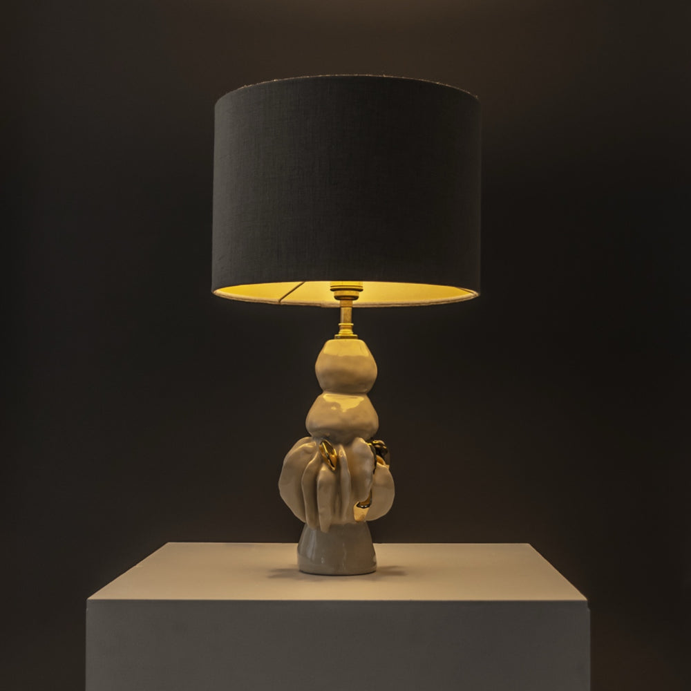 Handcrafted stoneware lamp glowing under a beige shade – sculptural base with organic form and optional linen cover. / Rankų darbo akmens masės šviestuvas su skulptūriniu pagrindu – šiltas apšvietimas tamsesniam interjerui.