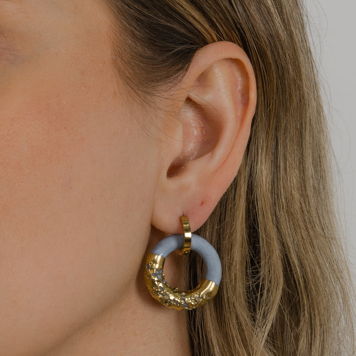 Close-up of an ear wearing a handmade gold and blue hoop earring against a neutral background