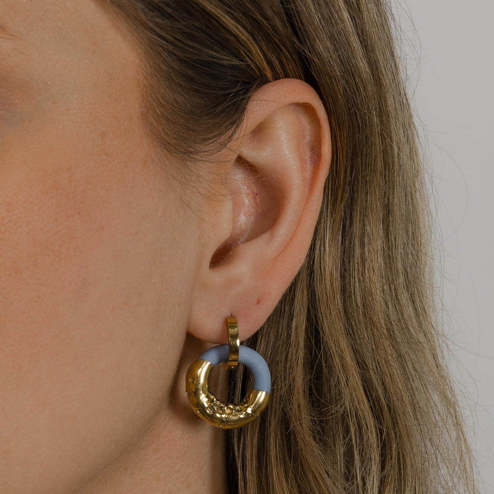 Ausis su auksiniu žiediniu auskaru ir rankų darbo porceliano pakabuku neutraliame fone, stambus planas