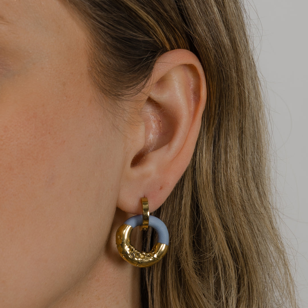 Ausis su auksiniu žiediniu auskaru ir rankų darbo porceliano pakabuku neutraliame fone, stambus planas