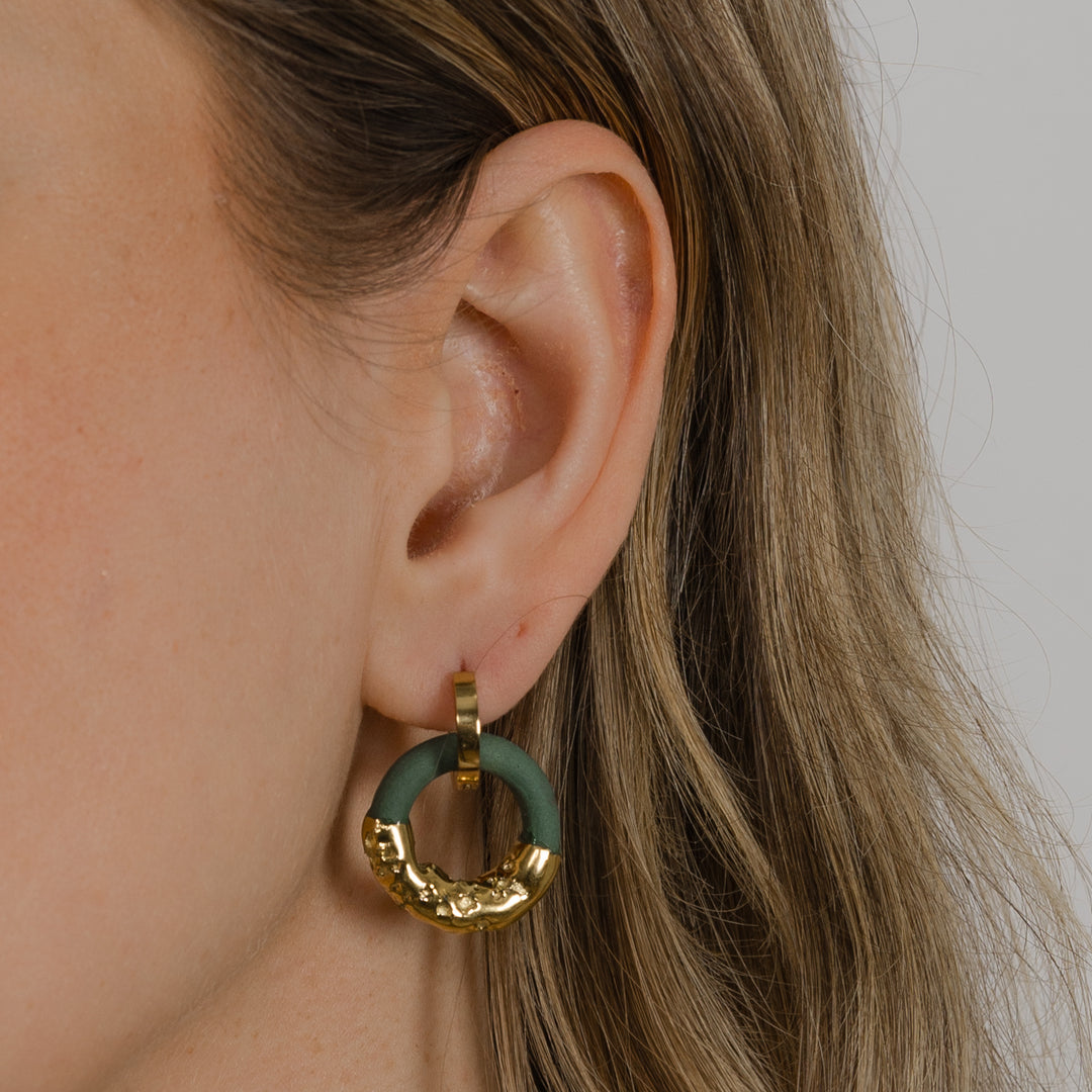 Ausis su auksiniu ir žaliu žiediniu auskaru neutraliame fone, stambus planas