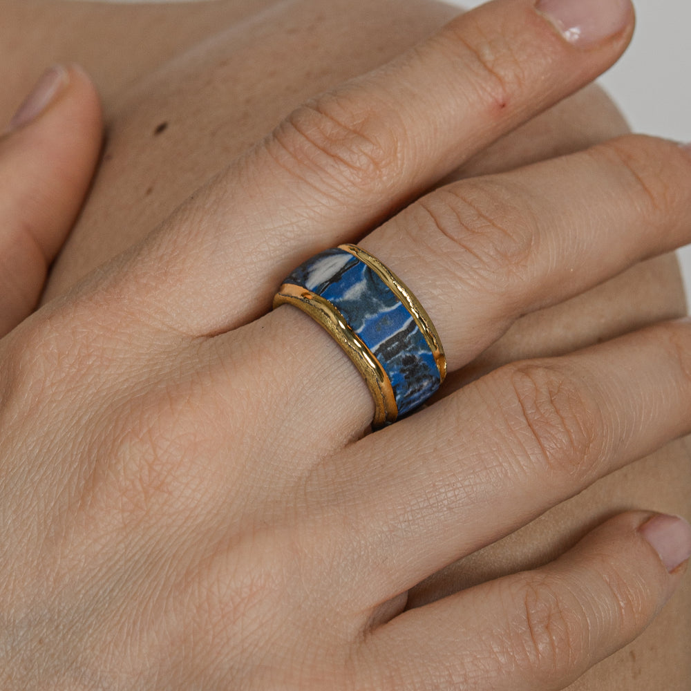 Hand wearing a gold and blue ring on a neutral background