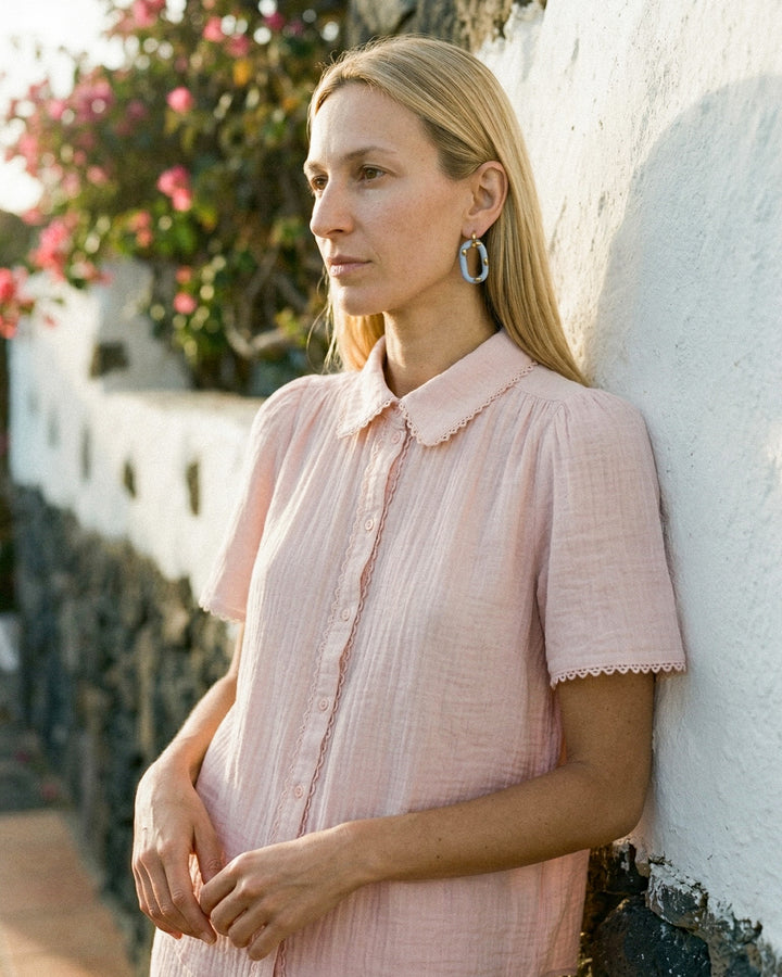 Moteris su rožine suknele ir rankų darbo porcelianiniu auskaru, kurį sukūrė freakyfoxx, atsiremia į baltą sieną, kurios fone – gėlės.