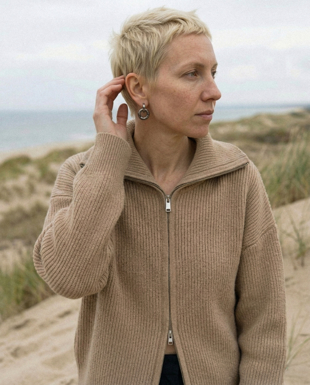 Moteris su rankų darbo keraminiais auskarais stovi smėlėtame paplūdimyje, fone – žolėtos Nidos kopos ir Baltijos jūra.