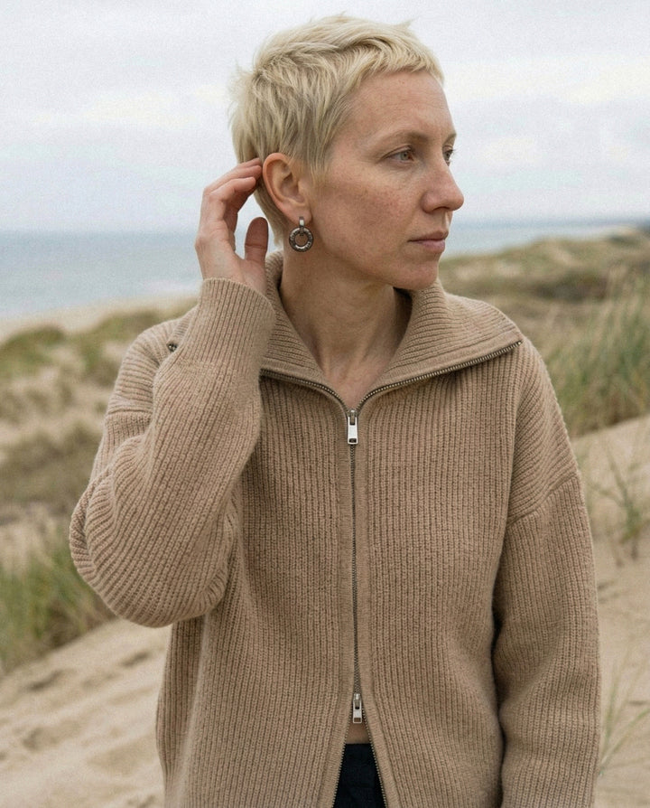 Moteris su rankų darbo keraminiais auskarais stovi smėlėtame paplūdimyje, fone – žolėtos Nidos kopos ir Baltijos jūra.