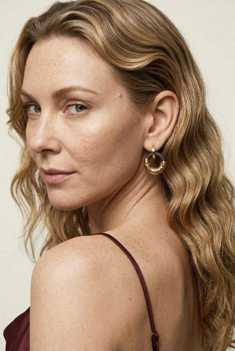 Moteris banguotais šviesiais plaukais, seginti auksiniais žiediniais auskarais neutraliame fone